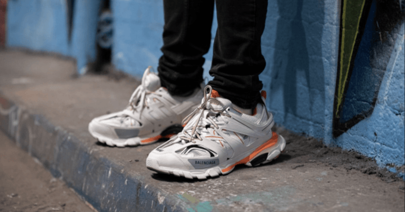 Man in a beige suit sitting on stone steps wearing grey, white and orange Balenciaga Track sneakers