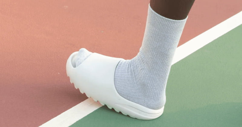 Two people wearing cream Yeezy Slides with black socks, sitting on a wooden floor