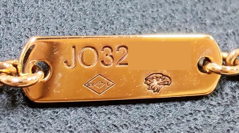 Achterkant van het gouden plaatje aan een Van Cleef & Arpels Alhambra ketting, gegraveerd met een serienummer dat begint met JO boven een meesterteken en de Franse adelaarskop, laatste cijfers afgedekt
